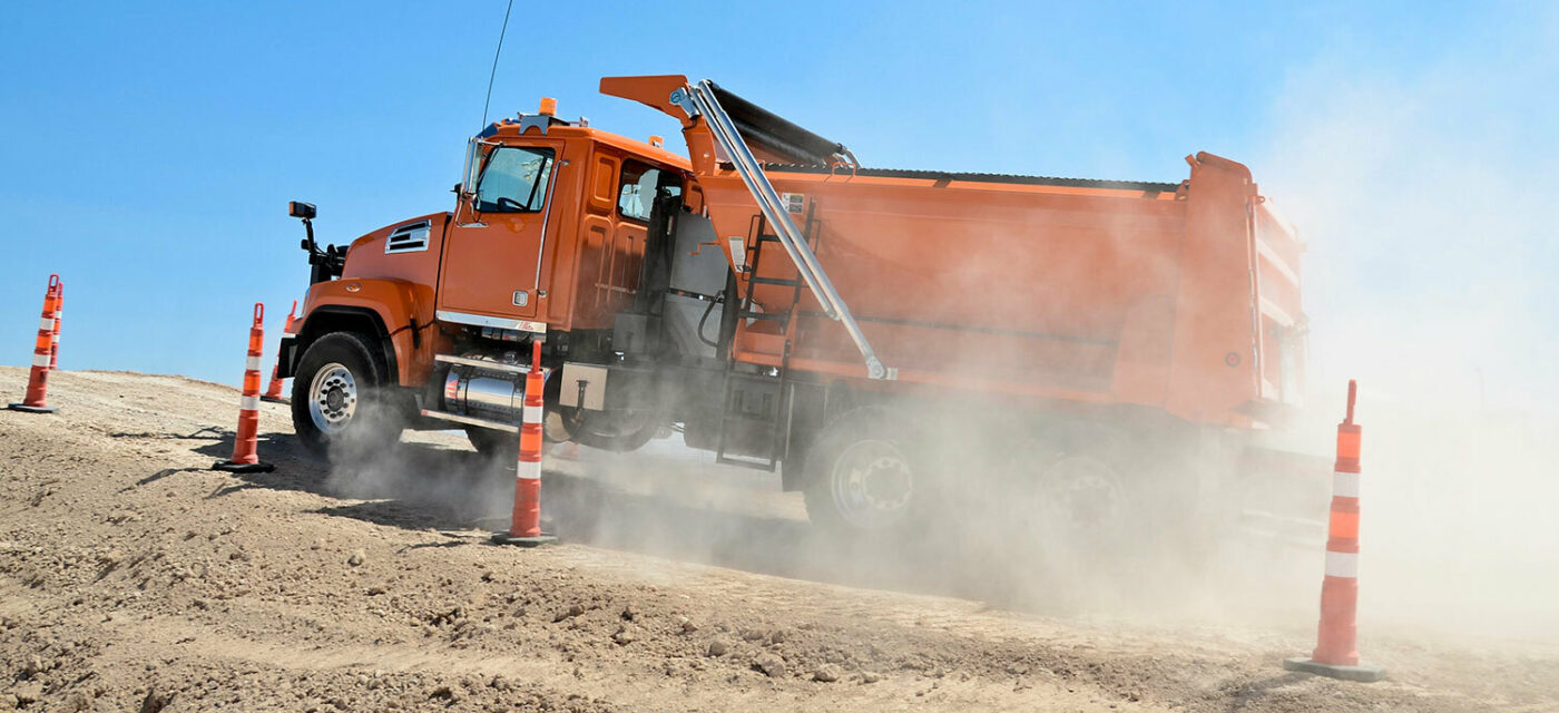 Orange dump truck driving up a dirt incline past caution cones