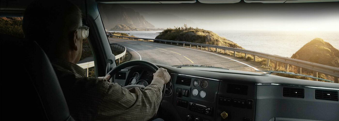 A photo taken from behind a driver in a semi cab looking out the windshield at a road and a view of the ocean