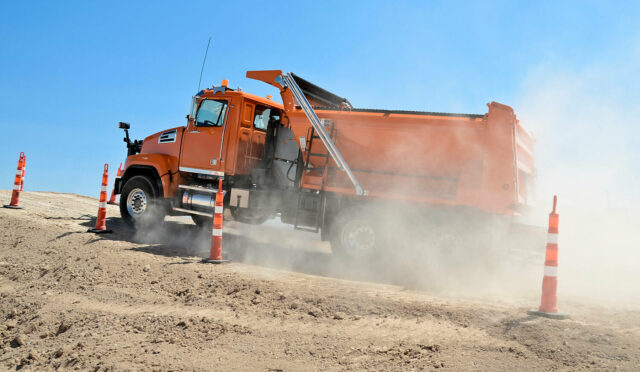 Orange dump truck driving up a dirt incline past caution cones