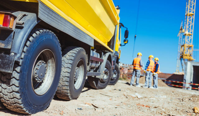 A low side angle photo of a yellow dump truck with three construction men to its right