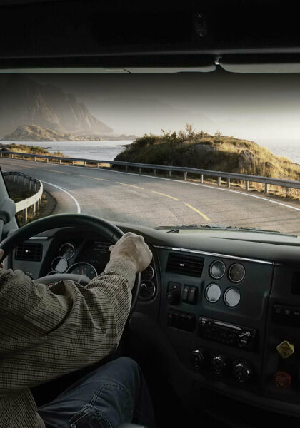 A photo taken from behind a driver in a semi cab looking out the windshield at a road and a view of the ocean