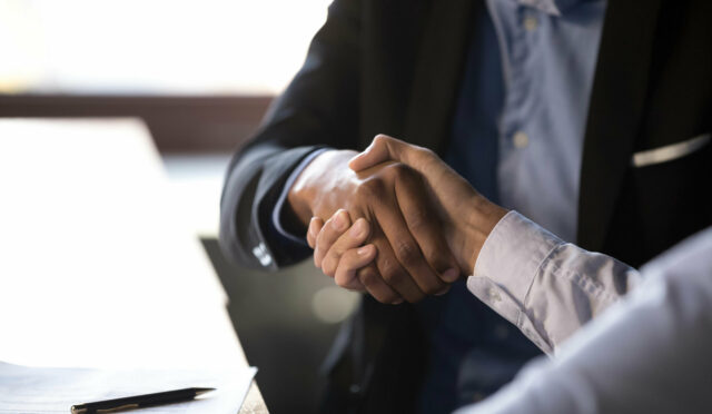 Two people shaking hands by document with a pen on top of it which is on top of a table