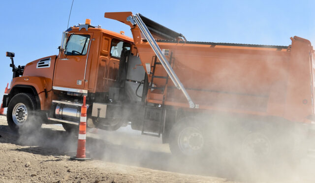 An orange dump truck with a lift axle driving up a dirt incline