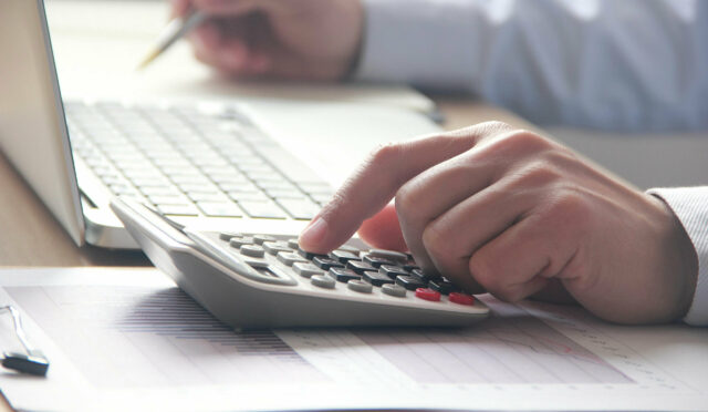a hand pushing buttons on an accounting calculator next to a laptop with another hand holding a pen on the other side of the laptop